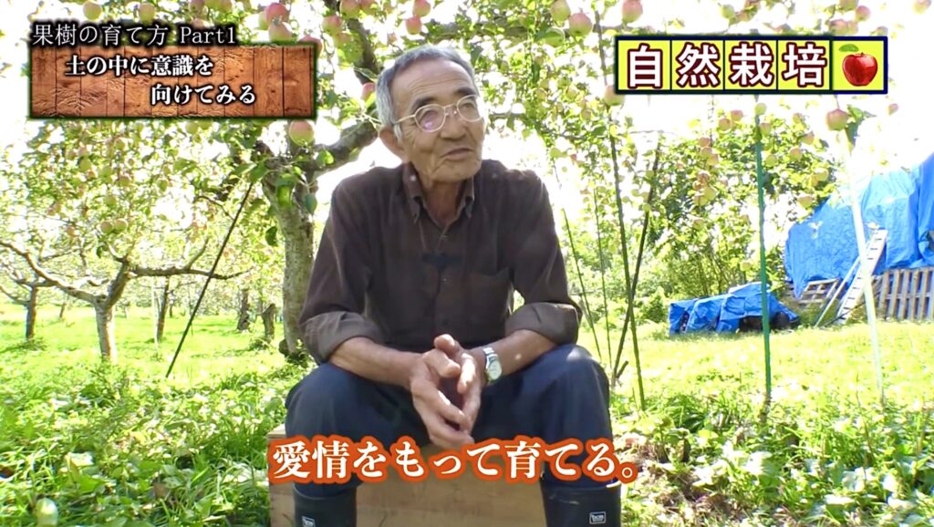 Screenshot - 奈良市 薬草|さくらハーブ JR奈良駅徒歩3分 Elderly man sits cross‑legged in an apple orchard, explaining natural cultivation as sunlight and fruit trees surround him (Japanese text overlays visible).